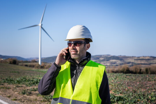 Engineer in protective helmet speaking on smartphone against wind turbines