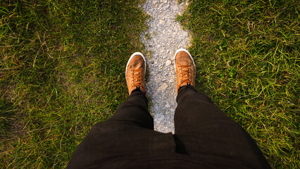 Top down to male feet on narrow path in grass, dressed in brown lace shows and black jeans | point of view | follow your path - obrazy, fototapety, plakaty