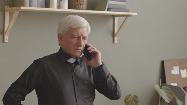 Senior grey haired priest in black shirt and clerical collar talking on phone while working from home office