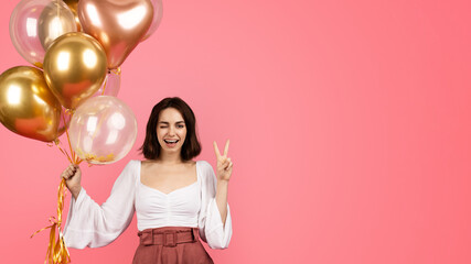 Happy young caucasian woman winks, hold inflatable balloons and peace sign with hand