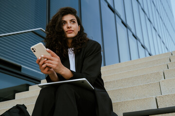 Young brunette businesswoman using cellphone and laptop outdoors