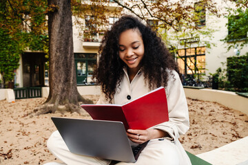 Fototapeta premium Smiling african girl doing homework with laptop while sitting outdoors