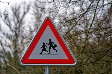 Red and white road warning sign at school house with triangle shape and trees in the background on a gray and cloudy winter day at City of Z&uuml;rich. Photo taken January 29th, 2023, Zurich, Switzerland.