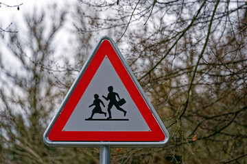 Red and white road warning sign at school house with triangle shape and trees in the background on a gray and cloudy winter day at City of Zürich. Photo taken January 29th, 2023, Zurich, Switzerland.
