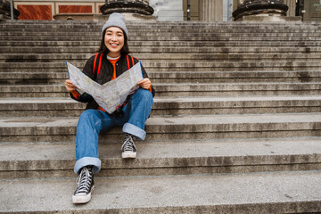 Positive asian girl tourist examining paper map while sitting on stairs