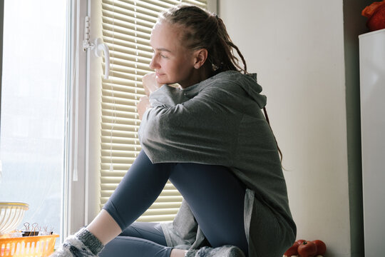 Young woman sitting on windowsill looking out the window with blinds