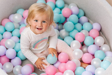 Fototapeta na wymiar Portrait close up Smiling blonde little baby kid girl lying on multi colored plastic balls in big dry paddling pool in playing centre. Having fun in playroom Leisure Activity.