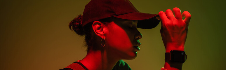 side view of african american woman in sweatshirt adjusting baseball cap on green with red light, banner.