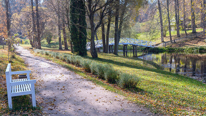white wooden bench near the path in the park near a small river with a wooden bridge