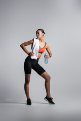 full length of african american woman in sports bra and bike shorts standing with sports bottle and towel on grey background.