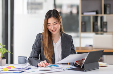 Young business Asian woman is sitting at table, working on laptop with graphs, charts, money and report financial concepts. Online marketing, education, analytics, e-commerce, business planning