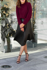 Young blond sexy girl woman in a romantic look in a red marsala winter cosy sweater and black eco leather skirt a in an urban city center new the glass wall with plants.