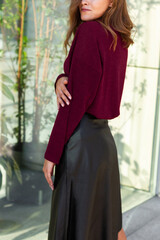 Fototapeta premium Young blond sexy girl woman in a romantic look in a red marsala winter cosy sweater and black eco leather skirt a in an urban city center new the glass wall with plants.