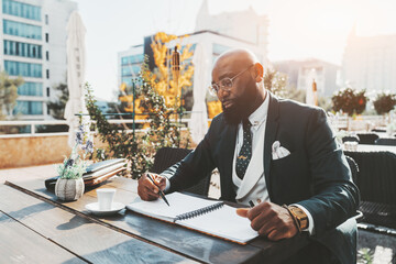 A confident mature hairless African American man entrepreneur in a tailored formal business suit and with a black beard is having a coffee break in an outdoor cafe while taking notes in his notebook