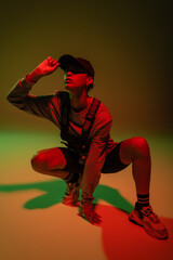 full length of african american woman in sweatshirt and vest sitting while adjusting baseball cap on green with red light.