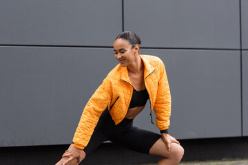 cheerful african american sportswoman in bike shorts and yellow puffer jacket exercising outside.