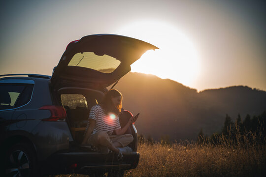 Young cheerful woman traveler sitting in open trunk of car enjoying sunset in mountains using smartphone
