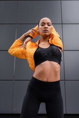 low angle view of african american sportswoman in sports bra and yellow puffer jacket warming up...
