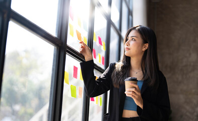 Young creative woman professional using sticky notes in glass wall to writing strategy business plan to development grow to success. Happy asian woman working at office.