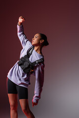 young african american woman in sweatshirt and bike shorts posing with raised hand on grey and purple.