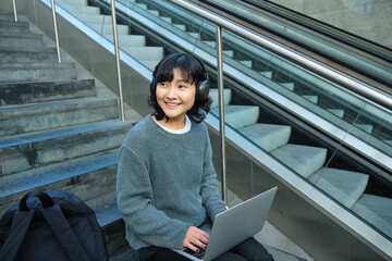 Portrait of beautiful young korean woman, student sits on stairs in public place, listens music in...