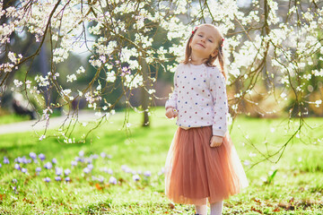 Adorable little girl enjoying nice and sunny spring day near apple tree in full bloom