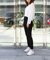 Young brunette sexy girl woman in a romantic look in a white winter cosy sweater and black pants and hat in an urban city center near the glass wall with plants.