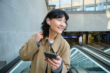 Fototapeta na wymiar Young brunette woman commutes, goes somewhere in city, stands on escalator and uses mobile phone, holds smartphone and smiles, listens music in wireless headphones