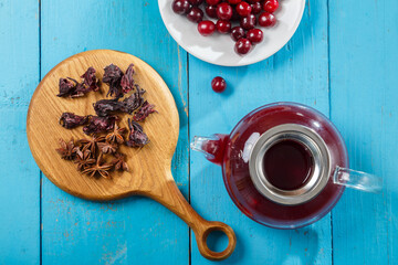 A teapot and a cup of hibiscus tea nearby on a blue table on a wooden board anise and cinnamon and hibiscus and cherry flowers in a plate. top view