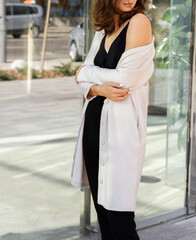 Fototapeta premium Young brunette sexy girl woman in a cosy look in a beige pale dress cardigan and black top and pants in an urban city center.