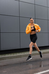 full length of cheerful african american sportswoman in bike shorts and yellow puffer jacket exercising outside.