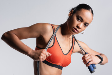 brunette african american sportswoman in red sports bra holding skipping rope isolated on grey.