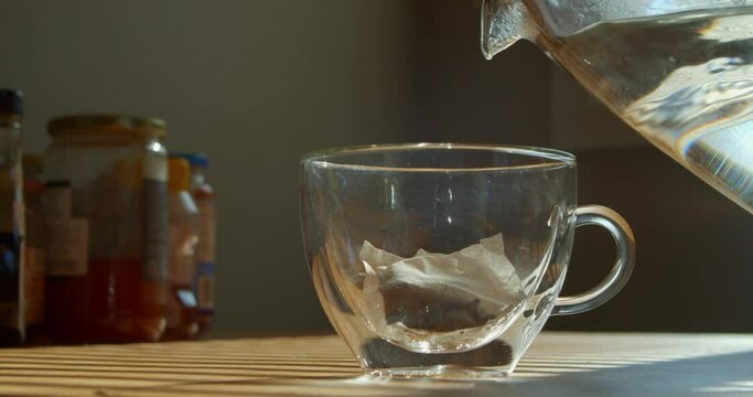 Making hot tea on the kitchen table. Boiling water is poured from the kettle into a cup with a tea bag. Transparent cup of freshly brewed tea on the kitchen table on a sunny day.
