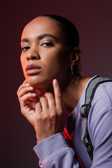 young african american woman in violet sweatshirt and vest looking at camera on dark purple.