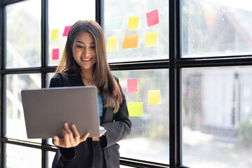 Asian freelance woman smiling paste post-it on the wall to record for work and using on laptop on wooden table at home. Entrepreneur woman working for her business at home. Business concept.