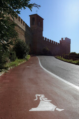 Funny pedestrian sidewalk in the medieval village of Gradara (Italy), with a middle age dressed lady over the white and red stripes of asphalt road, and village towers and walls on background - 566330066