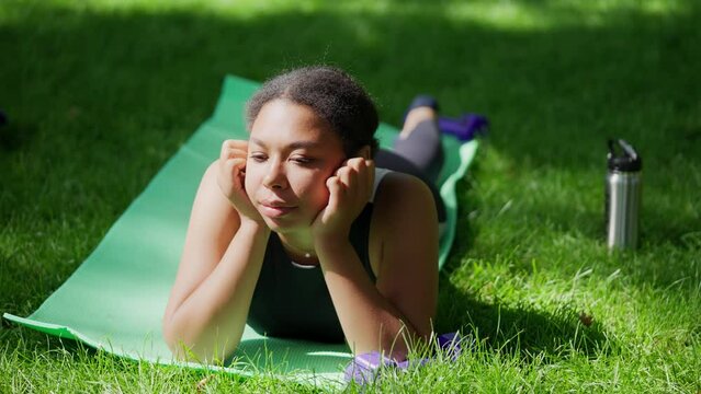 Dreamy African American woman resting on fitness mat in park, taking a break