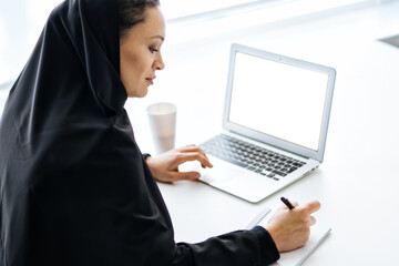 Beautiful woman with abaya dress working on her computer. Middle aged female employee at work in a...