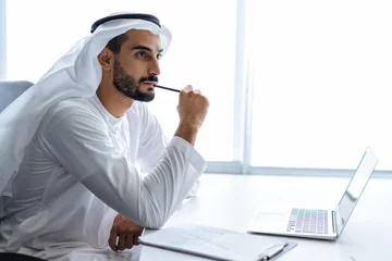 Foto auf Glas handsome man with dish dasha working in his business office of Dubai. Portraits of a successful businessman in traditional emirates white dress.  © oneinchpunch