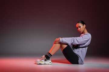 full length of african american woman in bike shorts and sweatshirt sitting on grey background with red light.