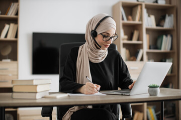 Attractive confident muslim business woman, office manager, wearing headset and hijab using laptop while making, writing financial report, using pen, on paper working indoors.