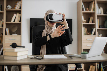 Muslim woman in headset touching virtual screen while sitting at desk with wireless laptop. Arab female freelancer in hijab using futuristic technology for work at home office.