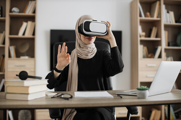 Young attractive muslim woman in hijab sitting at office and using VR headset and laptop for remote work Concept of people, technologies and freelance.