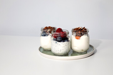 Yoghurts in glass jars with blueberries, raspberries and chia seeds and granola on a green plate on a white background