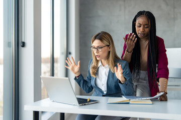 Two multiethnic business women in suit at office desk feeling disappointed, frustrated and sad over...