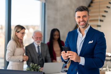 Happy confident handsome wealthy businessman leader holding mobile phone looking at camera standing in modern office for waist up shot photo. Diverse business team defocused in background.