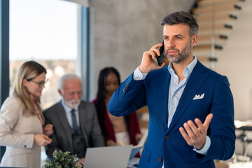 Serious millennial business man having a conversation on cell phone while standing in office fully focused to hear other side. Defocused diverse business team working in background.