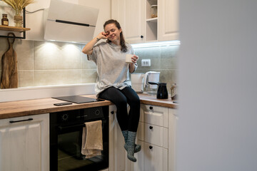 Woman sitting in kitchen in the morning holding mug of coffee rubbing her eyes and stretching her body and smiling.