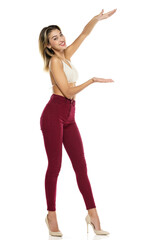 Smiling Woman In Burgundy Jeans And Top Presenting Something On her Empty Palm On A White Background