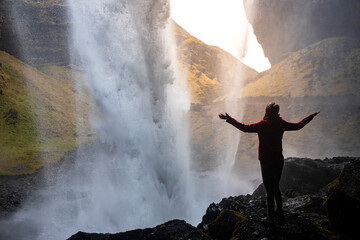 A girl in a pink jacket with arms outstretched standing in a cave on the rocks behind the powerful Kvernufoss waterfall, Iceland